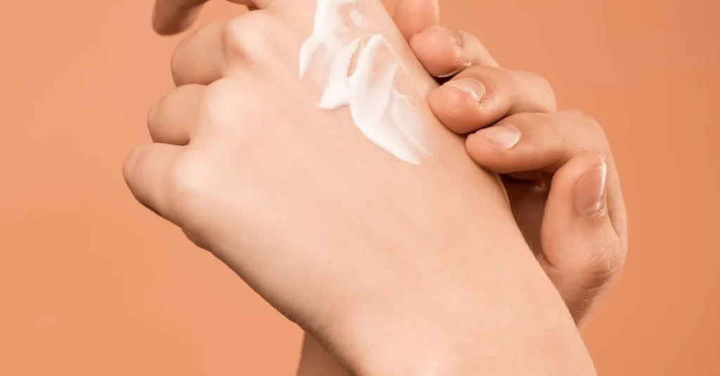 Close-up image of a person's hands applying white moisturizing cream on the back of another person’s hand against a soft peach background.